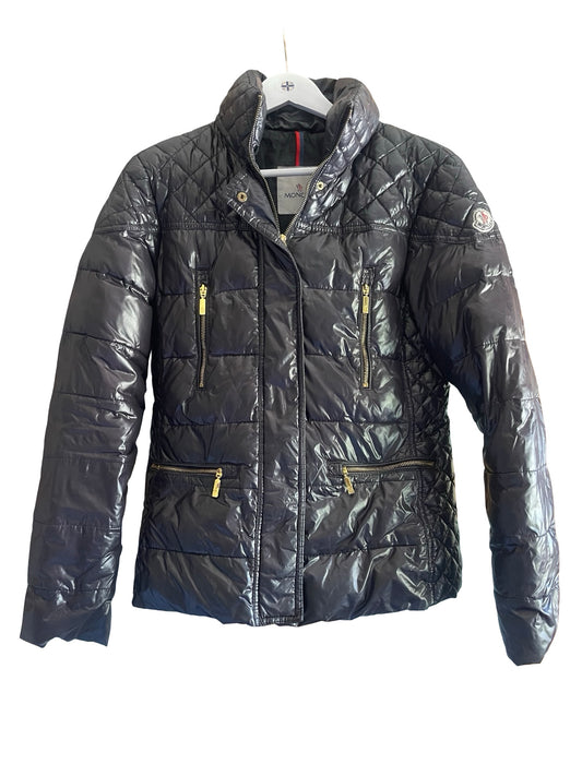 A black quilted puffer jacket with gold-tone zippers, hanging on a white hanger against a plain white background.