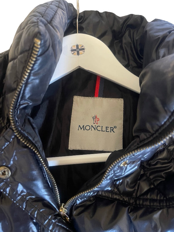 A close-up of a black Moncler puffer jacket on a white hanger, showing the brand's signature red and green stripe inside the collar and the Moncler logo patch on the hanger.