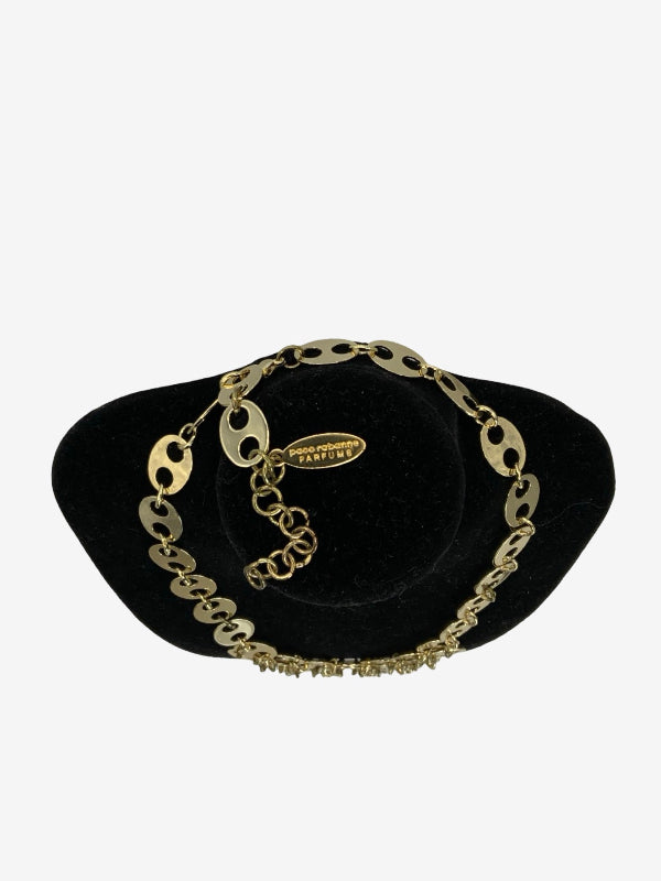 A gold-toned link chain bracelet with oval links and a small charm, displayed on a black velvet jewelry stand against a light gray background.