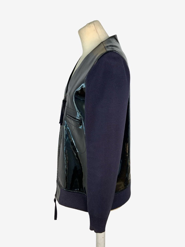A side-profile view of a long-sleeve knit sweater vest with glossy black patent leather panels, displayed on a mannequin torso against a light gray background.