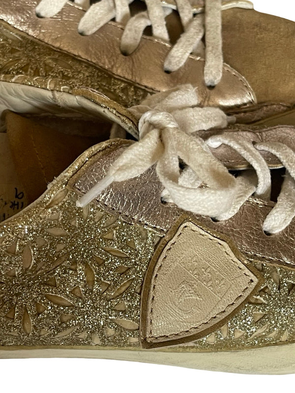 Close-up of a metallic gold sneaker with textured glittery detailing, white laces, and a shield-shaped logo patch on the side.