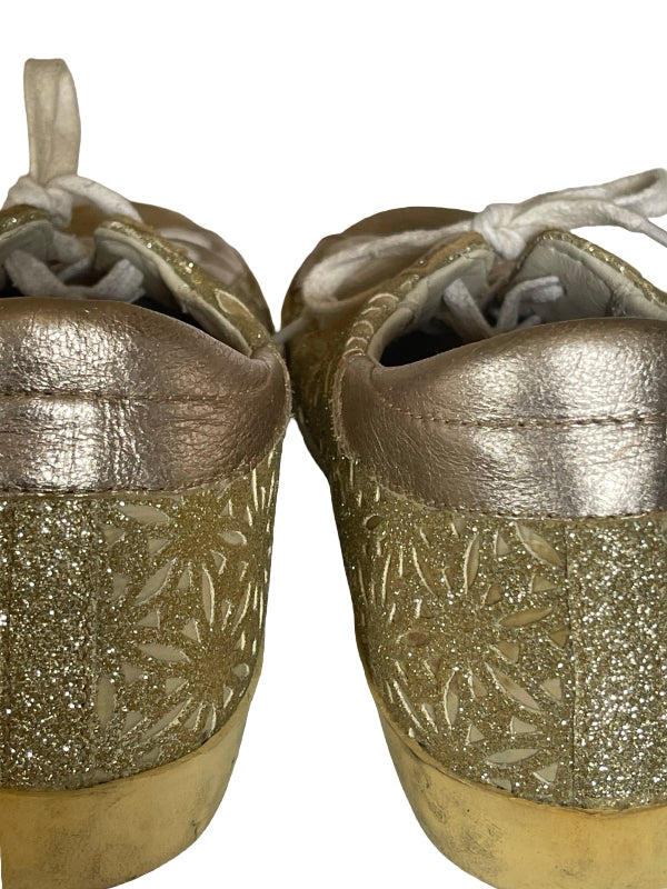 A pair of metallic gold low-top sneakers with glittery gold floral cutout patterns on the sides, white laces, and a gold sole, shown against a plain white background.