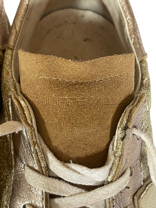 Close-up of a metallic gold sneaker showing the interior, featuring a tan suede tongue  with the brand name 'Philippe Model' embossed, white laces, and a white background.