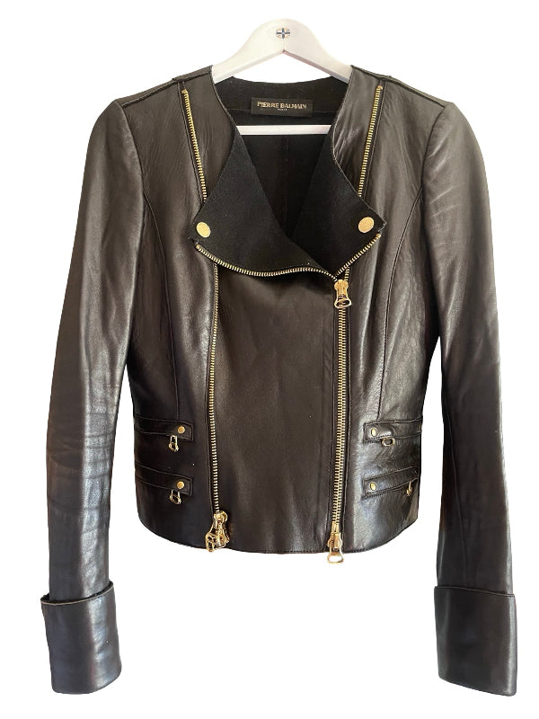 A black leather moto jacket with gold-tone hardware, hanging on a white hanger against a plain white background. The brand name 'PIERRE BALMAIN' is visible on the inner neck label.