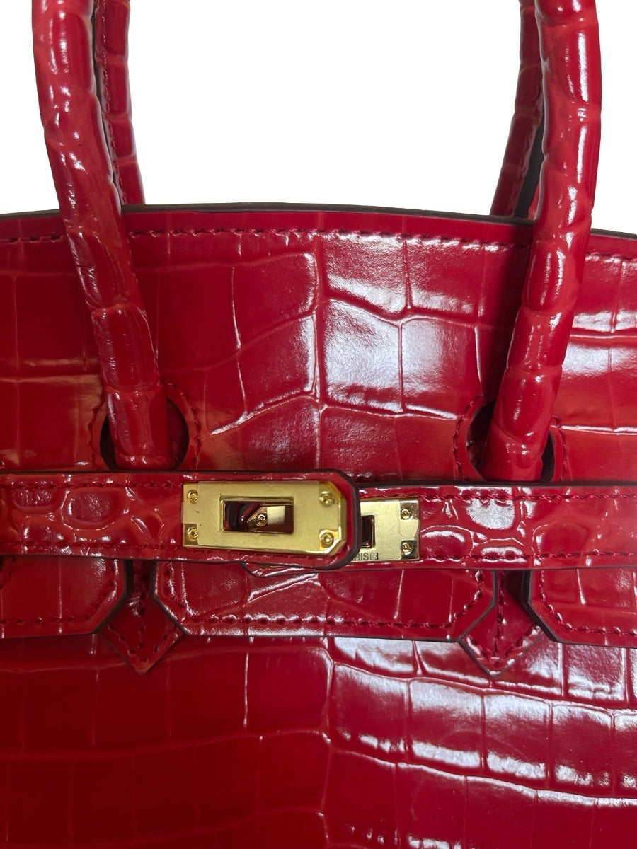 A close-up of a glossy red crocodile-embossed leather handbag with gold-tone hardware and top handles against a white background.