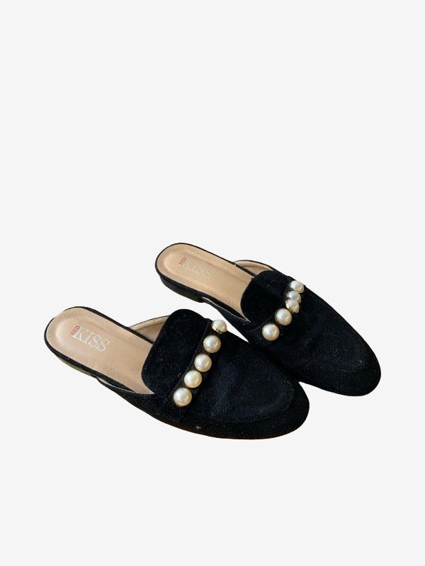 A pair of black velvet mule flats decorated with a row of white faux pearls across the top, shown on a light gray background.