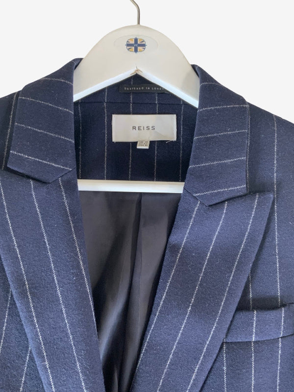 A close-up of a navy blue pinstripe blazer hanging on a white hanger with a small flag emblem. The brand label 'REISS' is visible inside the collar.