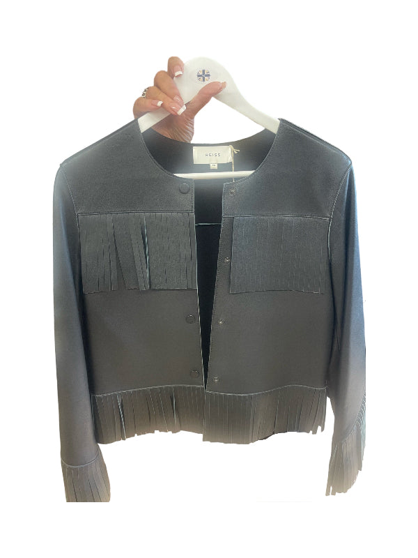A black fringed jacket with long sleeves and button-front chest pockets, held on a white hanger by a hand against a plain white background.