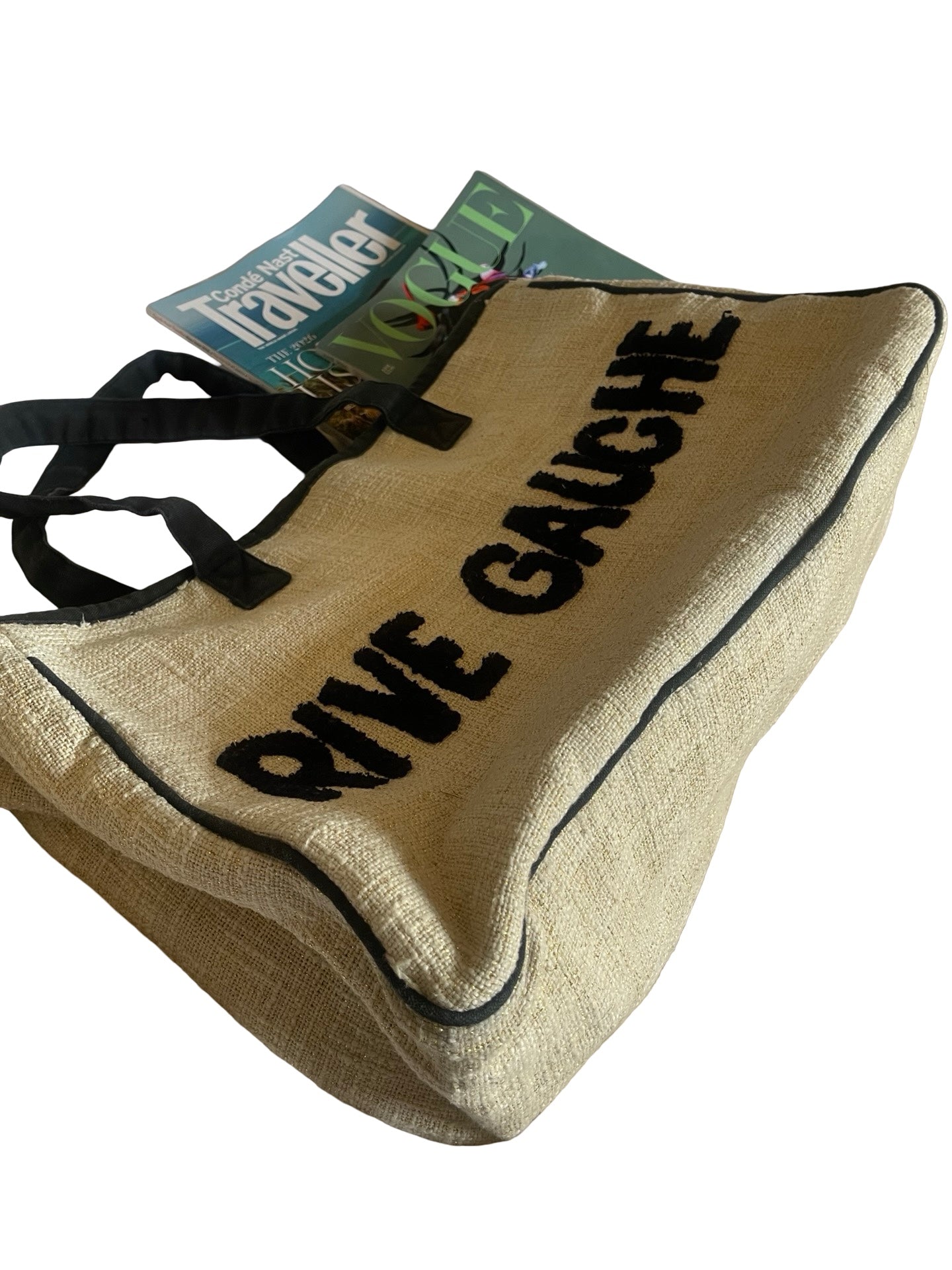 A natural jute tote bag with black trim and black handles, featuring the words 'RIVE GAUCHE' printed in bold black letters. A magazine is tucked inside the bag, which is photographed on a clean white background.