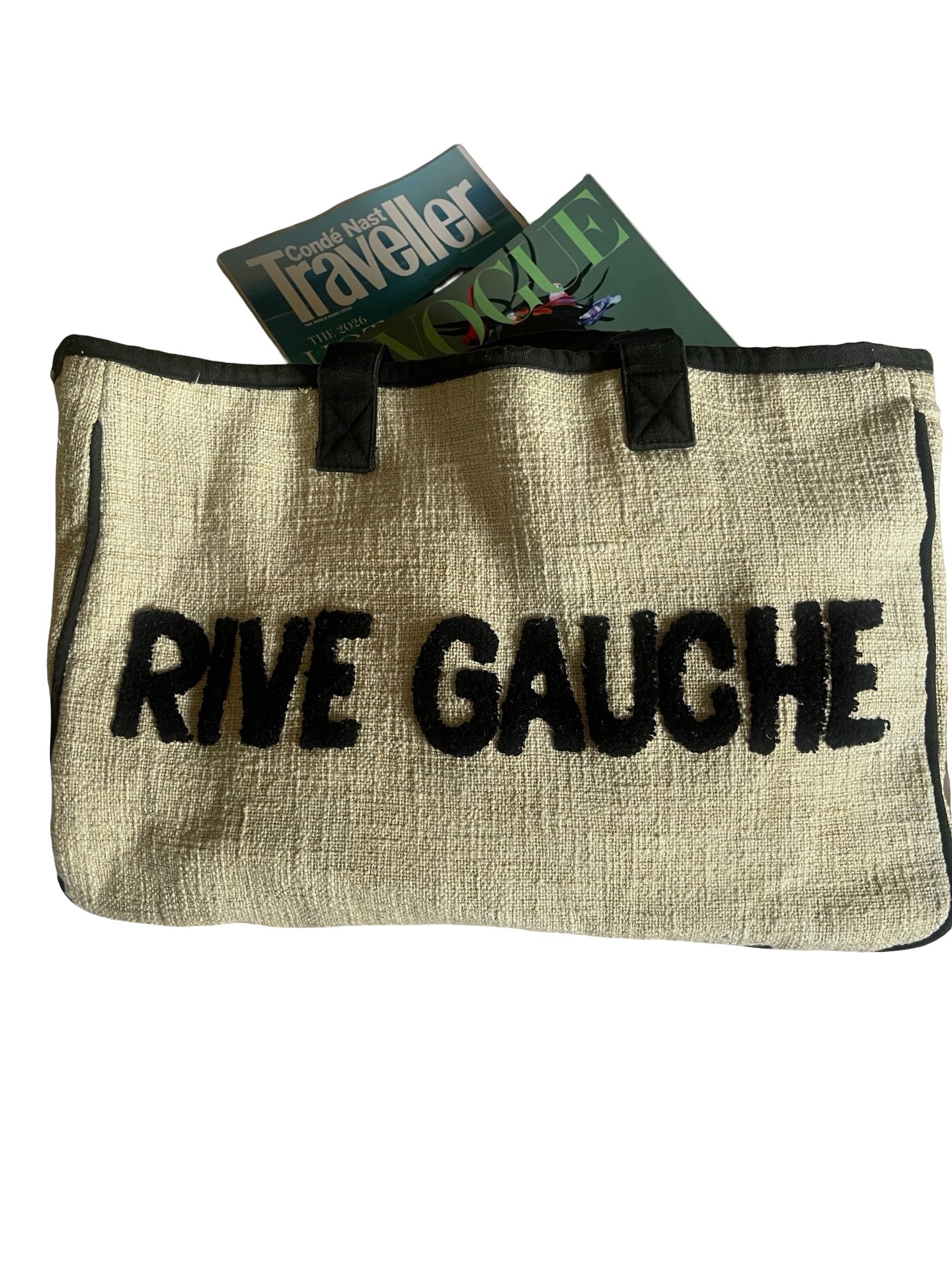 A beige linen tote bag with black trim and handles, featuring the words 'RIVE GAUCHE' printed in bold black letters across the front. Two magazines are tucked into the top opening of the bag against a plain white background.