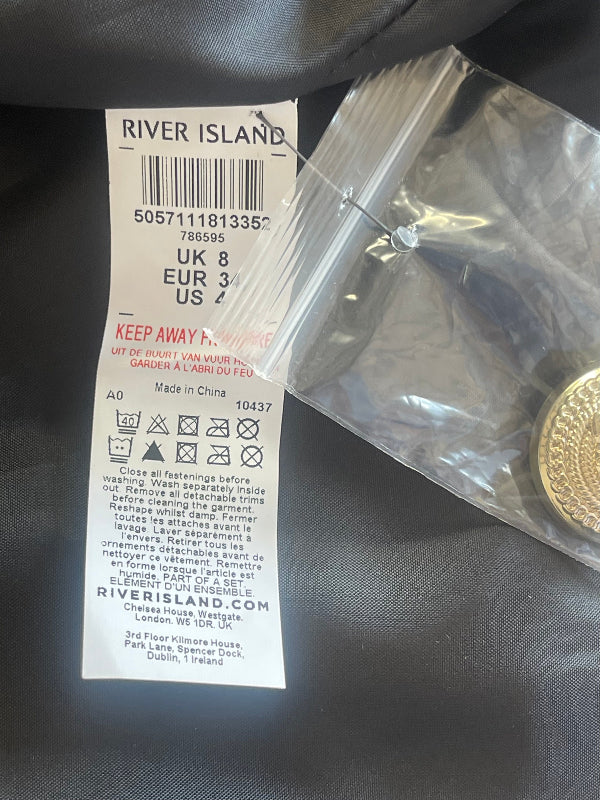 A close-up of a River Island product tag attached to a clear plastic bag containing a round gold-toned spare button, placed on a dark fabric surface