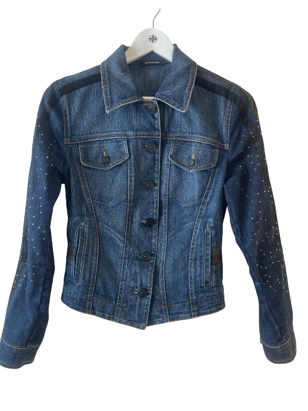 A blue denim jacket with black shoulder panels and silver rhinestone embellishments on the sleeves, hanging on a white hanger against a white background.