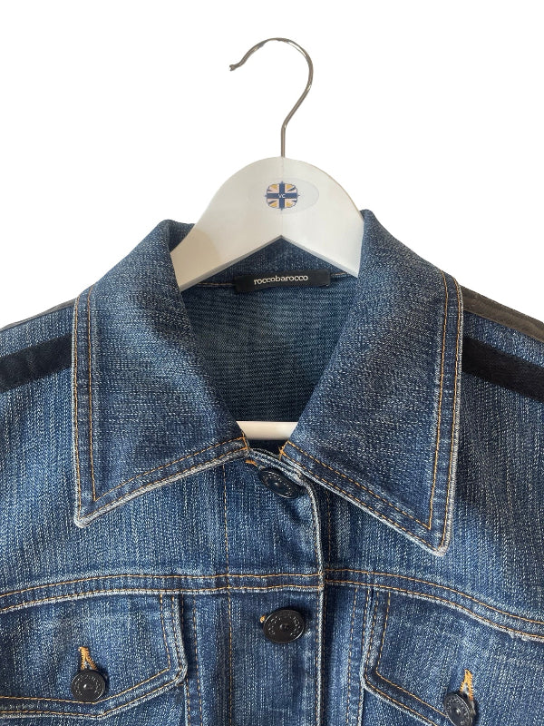 A close-up of a blue denim jacket hanging on a white hanger with a small flag logo. The jacket features contrast black shoulder panels, double-stitched details, and dark buttons.