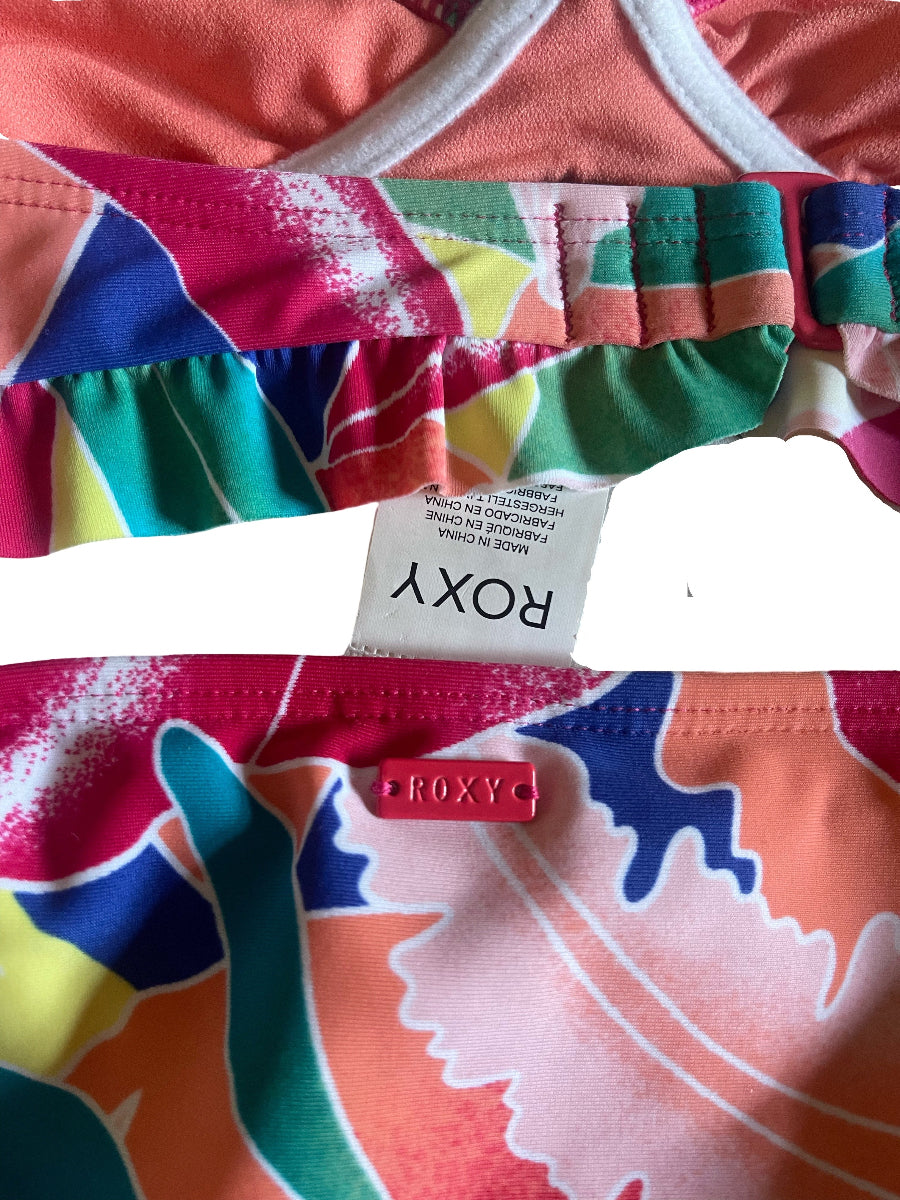 Close-up of a colorful patterned bikini bottom with a white Roxy brand tag visible at the center, showing a coral, pink, teal, green, yellow, and blue abstract print.
