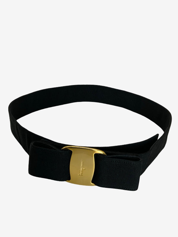 A black wide-gauge fabric belt with a large bow detail and a polished gold-tone buckle, shown on a light gray background.