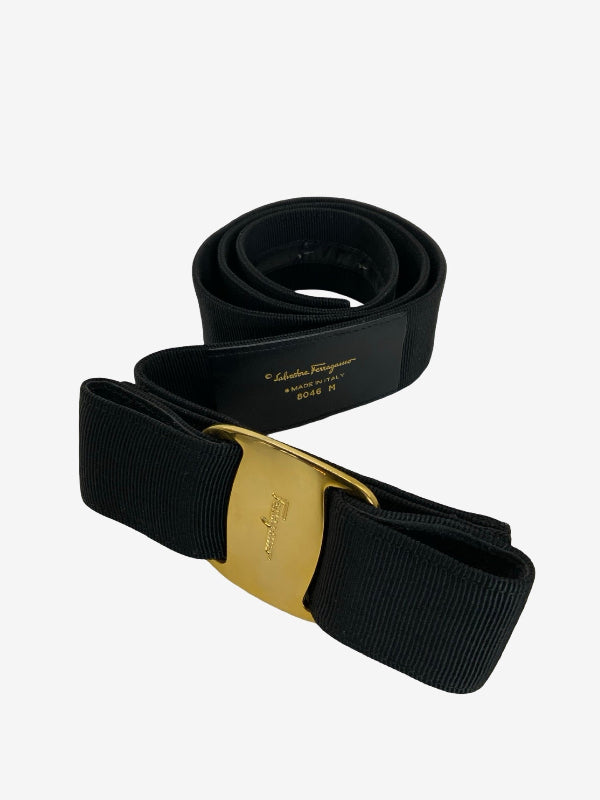 A black wide web belt with a gold-tone buckle, coiled on a light gray background. The buckle features the embossed logo of Celine's Salviatore design, and the interior tag reads 'salvatore ferragamo made in  ITALY M'.