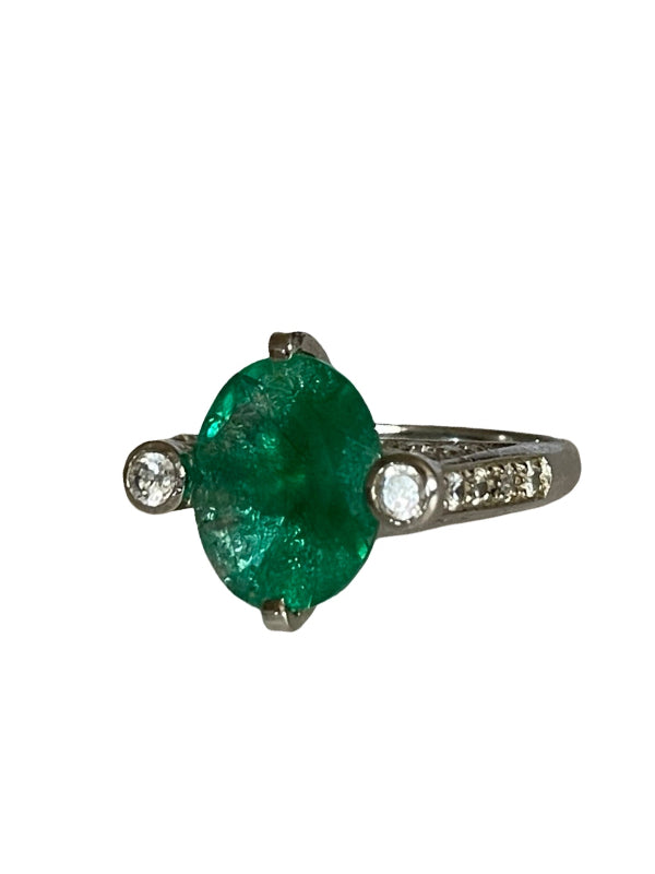 An oval-cut emerald green gemstone ring set in a silver-colored metal band with small clear accent stones on either side of the band, shown against a plain white background.
