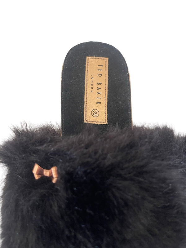 Close-up of a black fluffy faux-fur slipper with a tan leather TED BAKER London brand patch on the insole, set against a plain white background.