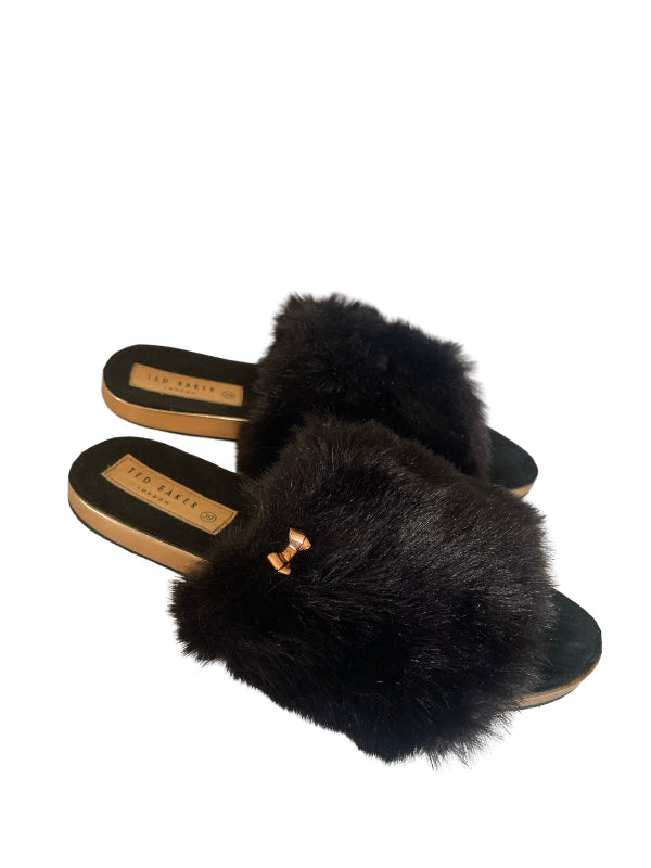 A pair of Ted Baker slide slippers featuring fluffy black fur uppers with a small gold bow detail, set against a plain white background.