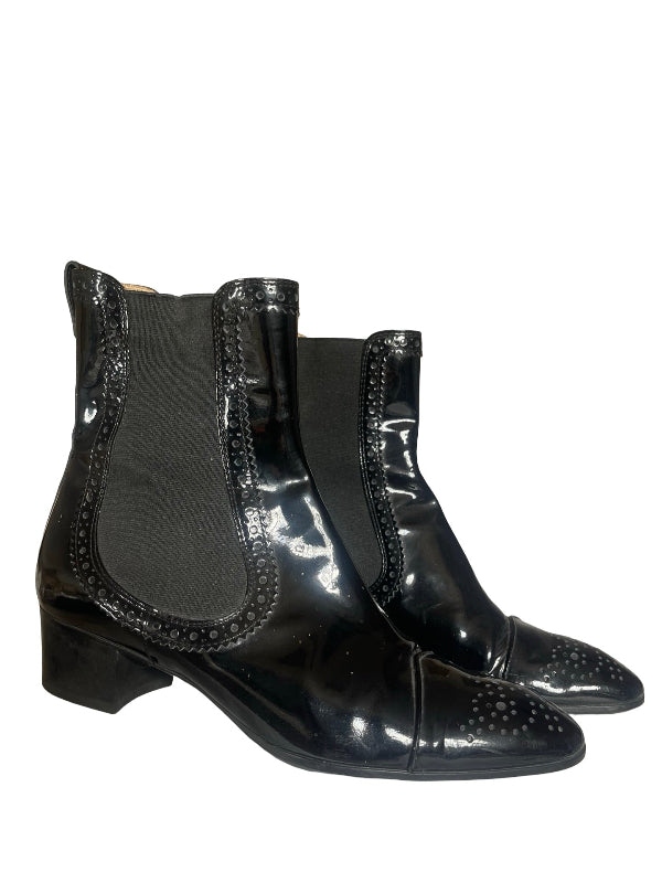 A pair of black ankle boots featuring glossy patent leather panels, elastic side panels, and decorative brogue detailing on the toe caps, set against a plain white background.