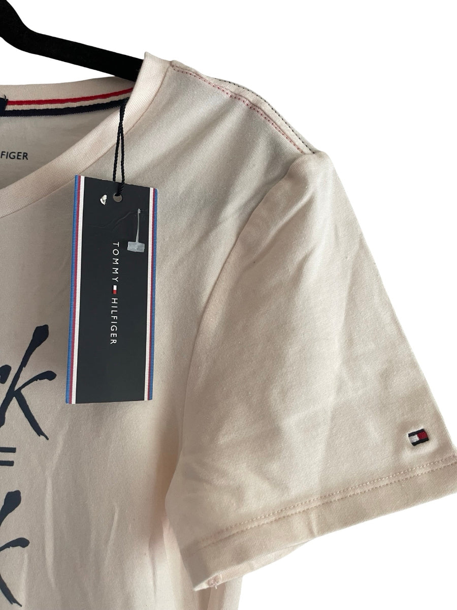 A close-up of a Tommy Hilfiger short-sleeve t-shirt in a light blush color, hanging on a black hanger with its original retail tag attached. The brand's signature red, white, and blue stripes are visible on the inner collar and the small flag logo on the sleeve cuff.