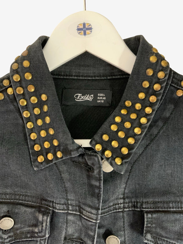 A close-up of a washed black denim jacket on a white hanger, featuring prominent gold-tone metal studs along the collar and button details.