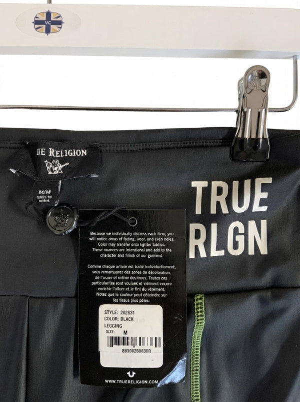 Close-up of a black True Religion garment clipped on a hanger, showing the True Religion label, hangtag, large white 'TRUE' text, and a green stitch detail