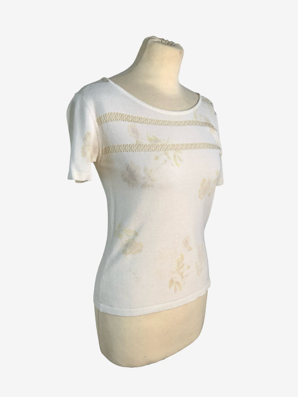 A white short-sleeve knit top with horizontal beige cut out stripes across the chest and subtle beige floral prints, displayed on a cream-colored mannequin torso against a light gray background.