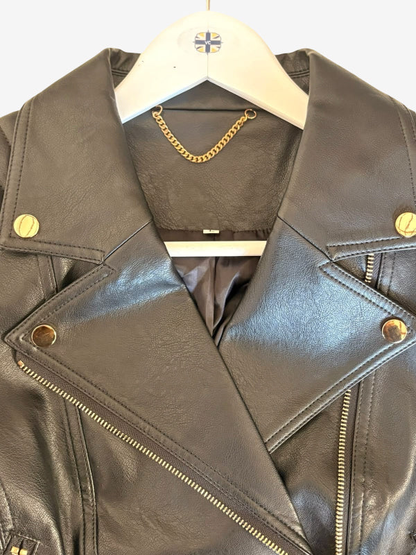 A close-up of a black faux leather motorcycle jacket hanging on a white hanger with a gold chain necklace inside the collar. The jacket features gold snap buttons and a diagonal zipper.