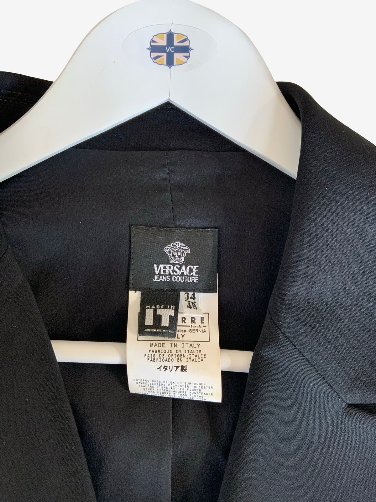 A close-up of a black Versace Jeans Couture blazer jacket hanging on a white hanger with a Versace logo patch. The inner neck label shows the brand 'Versace Jeans Couture' and size information.