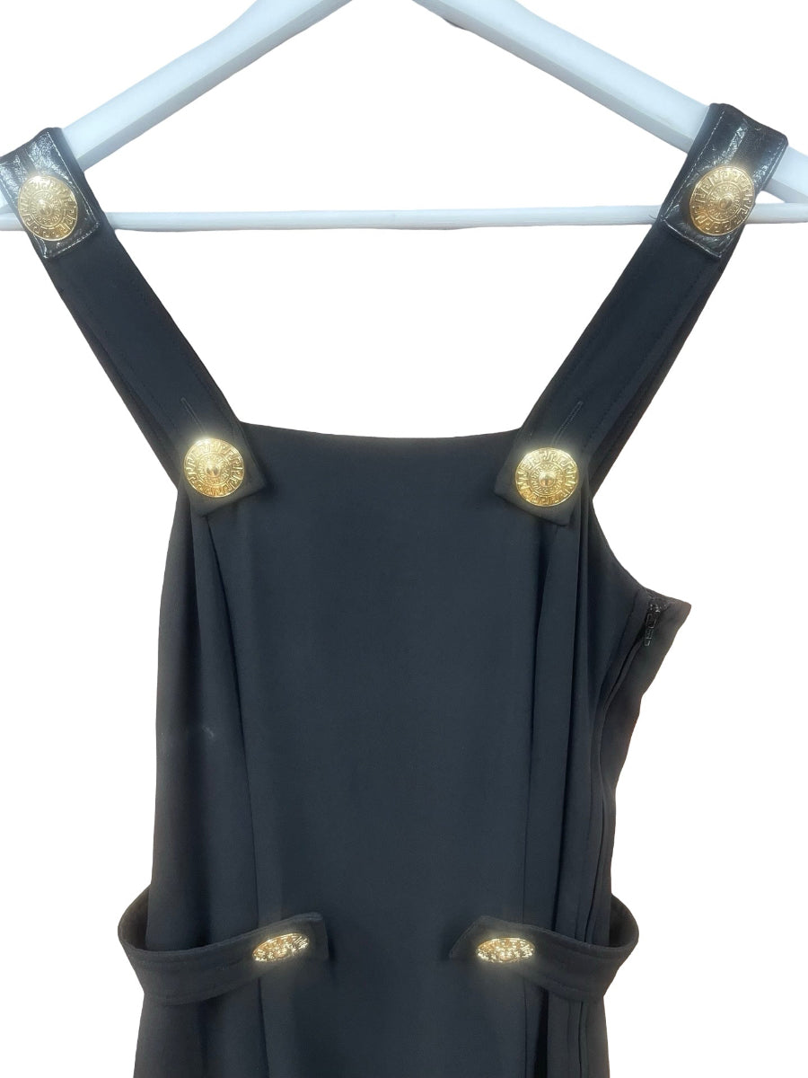 A black sleeveless dress or pinafore-style garment hanging on a white hanger against a white background, featuring wide shoulder straps and waist straps adorned with prominent gold-tone decorative buttons.