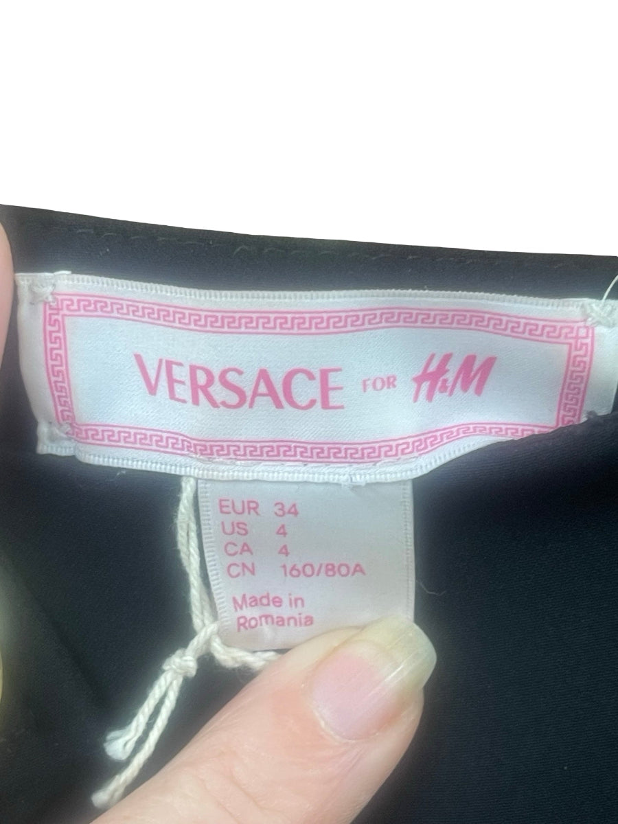 Close-up of a Versace for H&M garment label attached to a black garment, with a finger holding the hem. The label shows pink branding and size information.