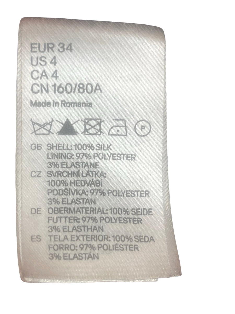 A close-up of a white fabric garment care label with black printed text and care symbols, including 'EUR 34', 'US 4', 'CA 4', and material information for shell, lining, and lining.