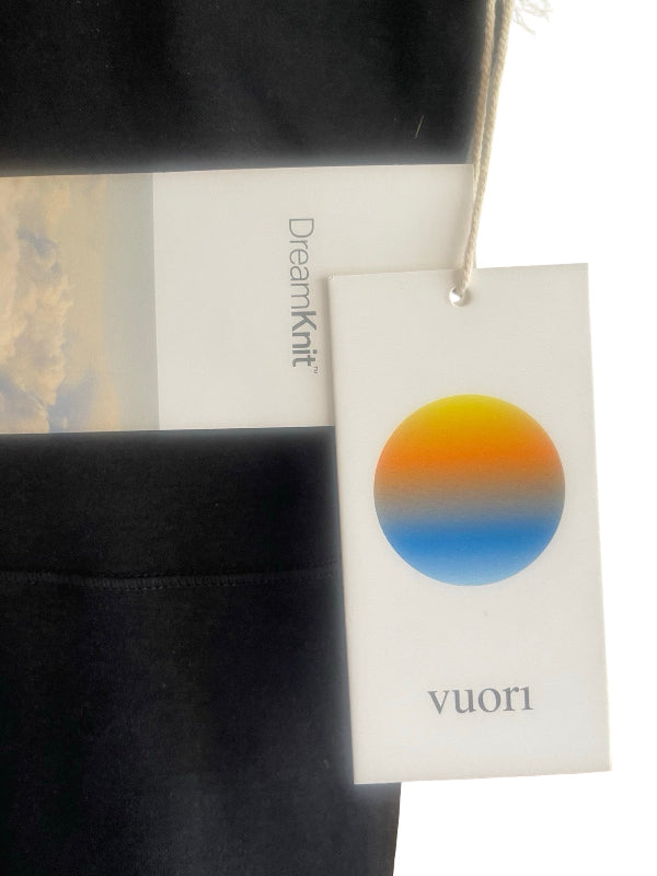 A close-up of a black garment with a white hangtag and a paper label. The tag has a circular gradient logo in yellow, orange, and blue, with the brand name 'vuori' printed at the bottom.