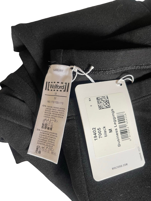 A close-up of folded black trousers or leggings with two tags attached: a white content  tag and a white tag showing the size and product details.
