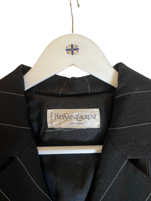 A black Yves Saint Laurent gabardine jacket with thin white pinstripes, hanging on a white wooden hanger with a Saint Laurent logo, showing the brand label inside.